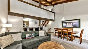 A modern living room with a sofa, coffee table, and dining area. A kitchen is visible in the background.