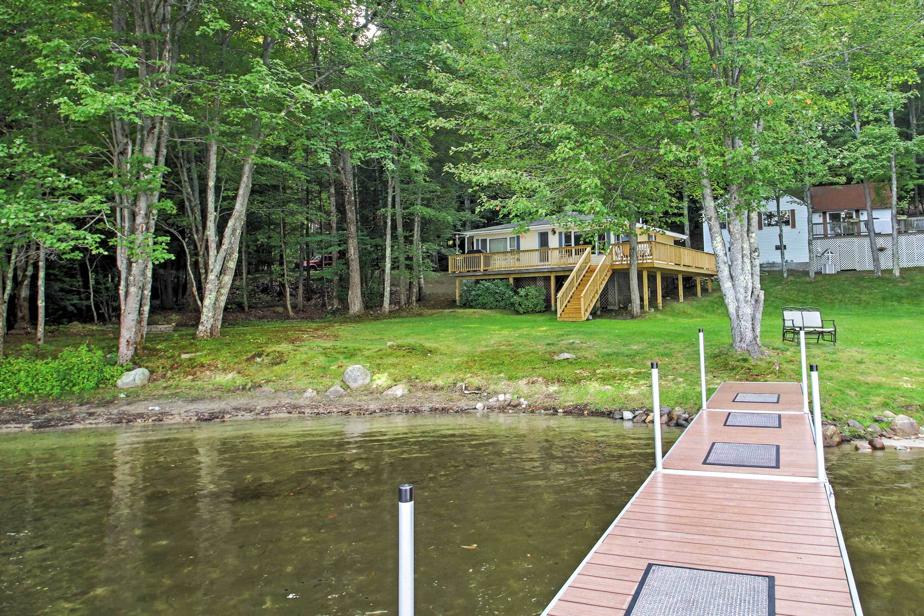 A wooden dock extending into a calm lake, surrounded by lush greenery and a house with a deck.