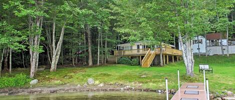 A wooden dock extending into a calm lake, surrounded by lush greenery and a house with a deck.