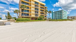 A multi-story hotel with balconies, a parking area, and a sandy beach in the foreground.