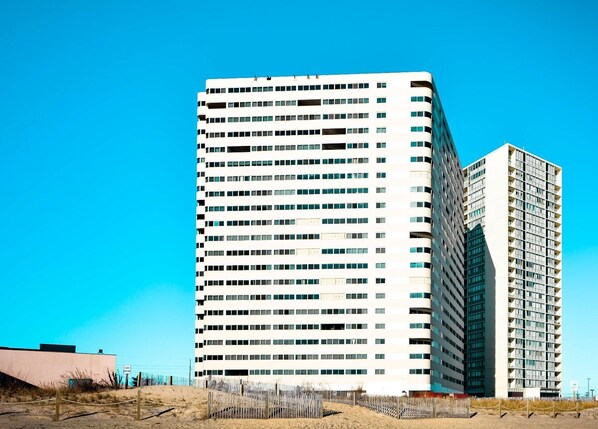 A tall, modern building with multiple floors and a flat roof, surrounded by a sandy area with a fence. - Kid-Friendly 3 Bedroom Oceanfront Condo with Enclosed Balconies and an Outdoor Pool Located Uptown and Mere Steps to the Beach! (Ocean City)