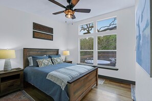 Un dormitorio con una cama de madera, dos lámparas de noche, un ventilador de techo y un ventanal con vistas a un árbol y una piscina.