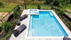Ein rechteckiger Swimmingpool mit Whirlpool, umgeben von einem schwarzen Metallzaun, und ausgestattet mit mehreren Liegestühlen.