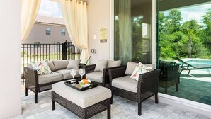 Eine sonnendurchflutete Terrasse mit Korbmöbeln, eine Glastür, die zum Pool führt, und ein Blick auf ein Haus im Hintergrund.