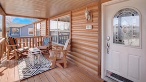 Una terraza de madera con sillas Adirondack y una pequeña mesa, una puerta blanca con un ventanal y el número '303' en la pared.