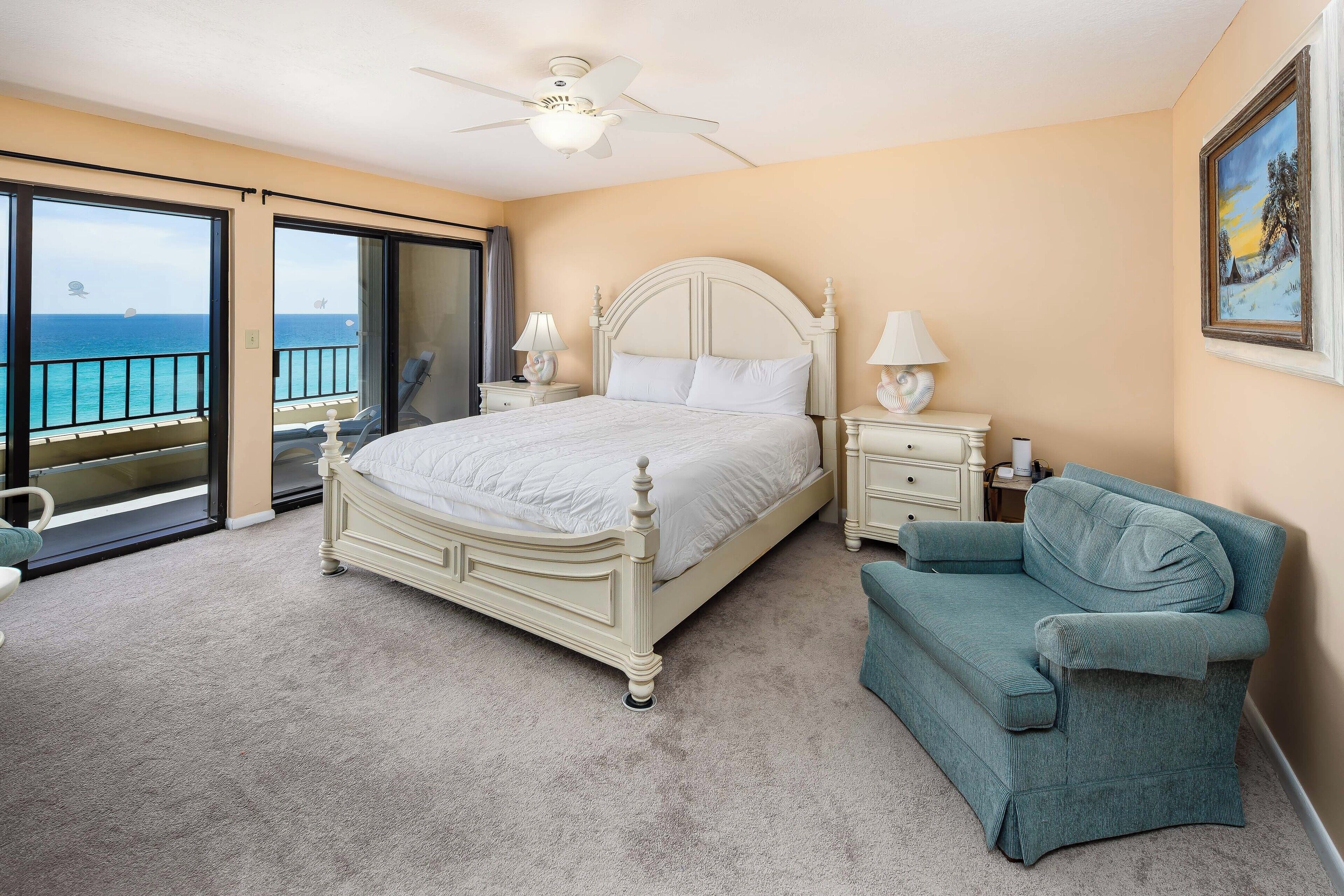 A bedroom with a large bed, a chair, a nightstand, and a view of the ocean through a sliding door.