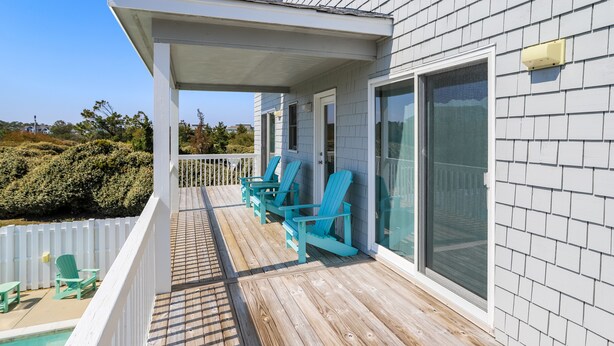 A covered deck with wooden flooring and a white railing, adjacent to a swimming pool and a house with large windows and a white fence.