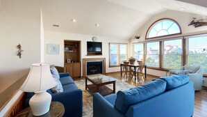 A spacious living room with a fireplace, blue sofas, a dining table with chairs, and large windows offering a view of the outside.