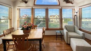 A dining area with a wooden table and chairs, a basket of flowers, and a view of houses through large windows.
