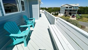 Eine Holzterrasse mit zwei türkisblauen Adirondack-Stühlen, einem weißen Geländer und Blick auf ein zweistöckiges Gebäude mit Garage und einem davor stehenden Auto.