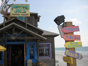 Un edificio de madera con un letrero que indica 'Pompano Joe's Seafood House' y varios letreros direccionales para 'Camisetas', 'Bar Tiki', 'Asientos en la terraza', 'Bar de playa' y 'Buena onda'.