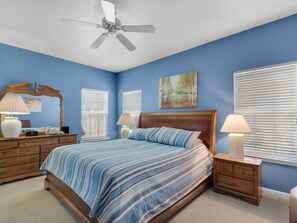 Un dormitorio con cama, mesitas de noche, cómoda, ventilador de techo y dos ventanas con persianas.