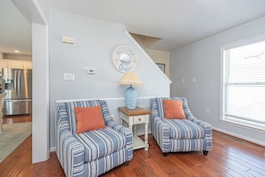 A living room with two striped armchairs, a small table with a lamp, a 'Welcome to the Ocean' sign, and a kitchen in the background.