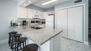 A modern kitchen with white cabinets, a granite countertop, and a double sink. There are four bar stools and a refrigerator. - ETW 1003: Sandy Toes - Where summer is a state of mind! (Fort Walton Beach)
