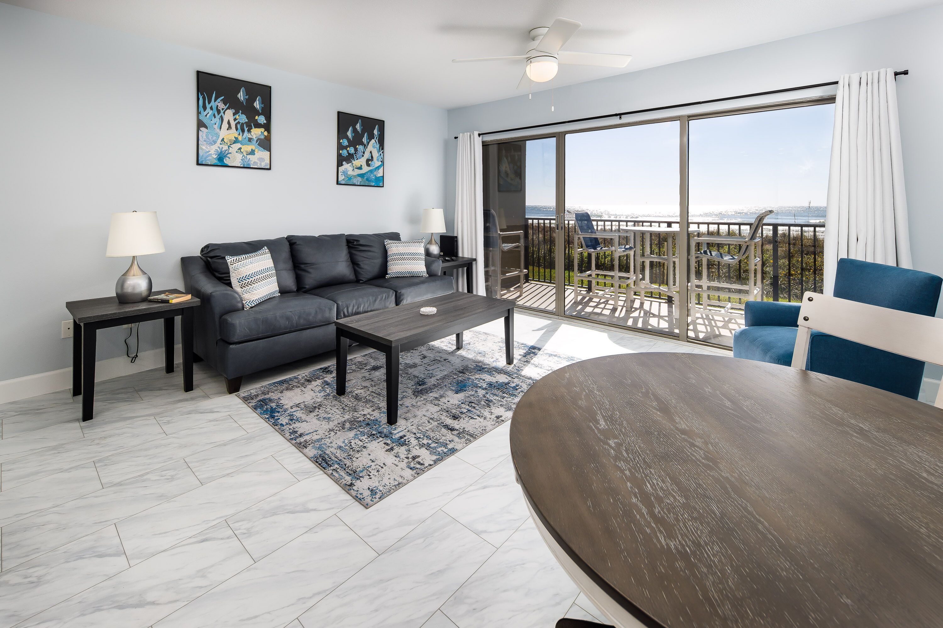 Un salon moderne avec un canapé gris, une table basse et un fauteuil bleu. Il y a un balcon avec vue sur les montagnes.