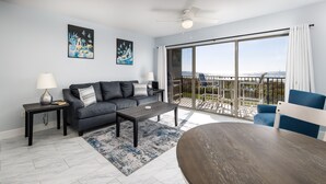 A modern living room with a grey sofa, a coffee table, and a blue chair. There is a balcony with a view of the mountains. - ETW 1003: Sandy Toes - Where summer is a state of mind! (Fort Walton Beach)