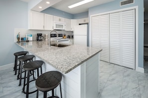 A modern kitchen with white cabinets, a granite countertop, and a double sink. There are four bar stools and a refrigerator. - ETW 1003: Sandy Toes - Where summer is a state of mind! (Fort Walton Beach)