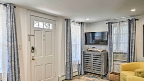 A living room with a white door, grey curtains, a television, a grey cabinet, and a yellow armchair. - Cozy Ocean Grove Beach Retreat: Walk to the Coast! (Ocean Grove)