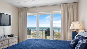 A hotel room with a large bed, a television, a dresser, and a view of the beach through the window.
