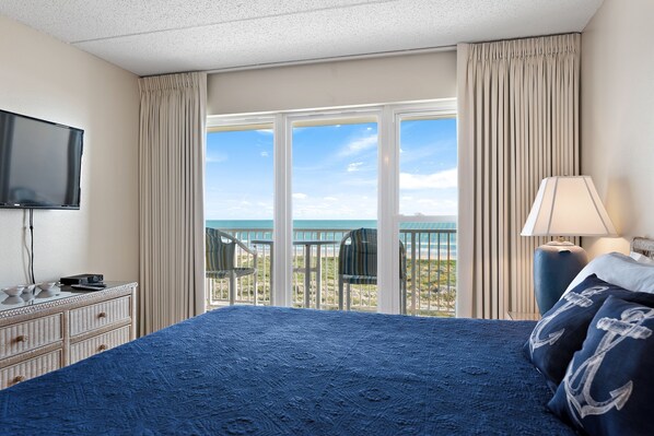 A hotel room with a large bed, a television, a dresser, and a view of the beach through the window.