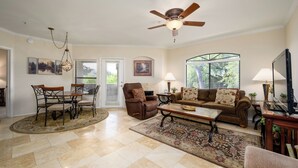 A living room with a dining area, a ceiling fan, a fireplace, and a large window with blinds.