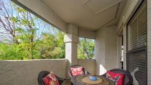 A covered outdoor seating area with wicker chairs and red cushions, a round mosaic table, and a view of trees and sky through a window.