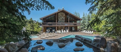 A lodge with a pool surrounded by rocks, a deck with lounge chairs, and a forested backdrop.