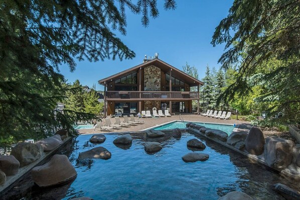 A lodge with a pool surrounded by rocks, a deck with lounge chairs, and a forested backdrop.