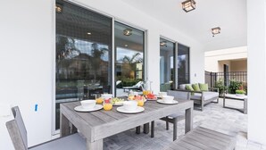 Un espace repas extérieur moderne, avec une table en bois dressée pour le petit-déjeuner, de grandes fenêtres et une vue sur les environs.