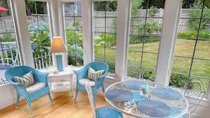 Habitación soleada con muebles de mimbre, una mesa de vidrio y vistas a un jardín a través de grandes ventanales.
