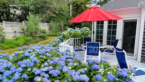 Un patio con una mesa blanca y sillas azules, rodeado de arbustos de hortensias azules.