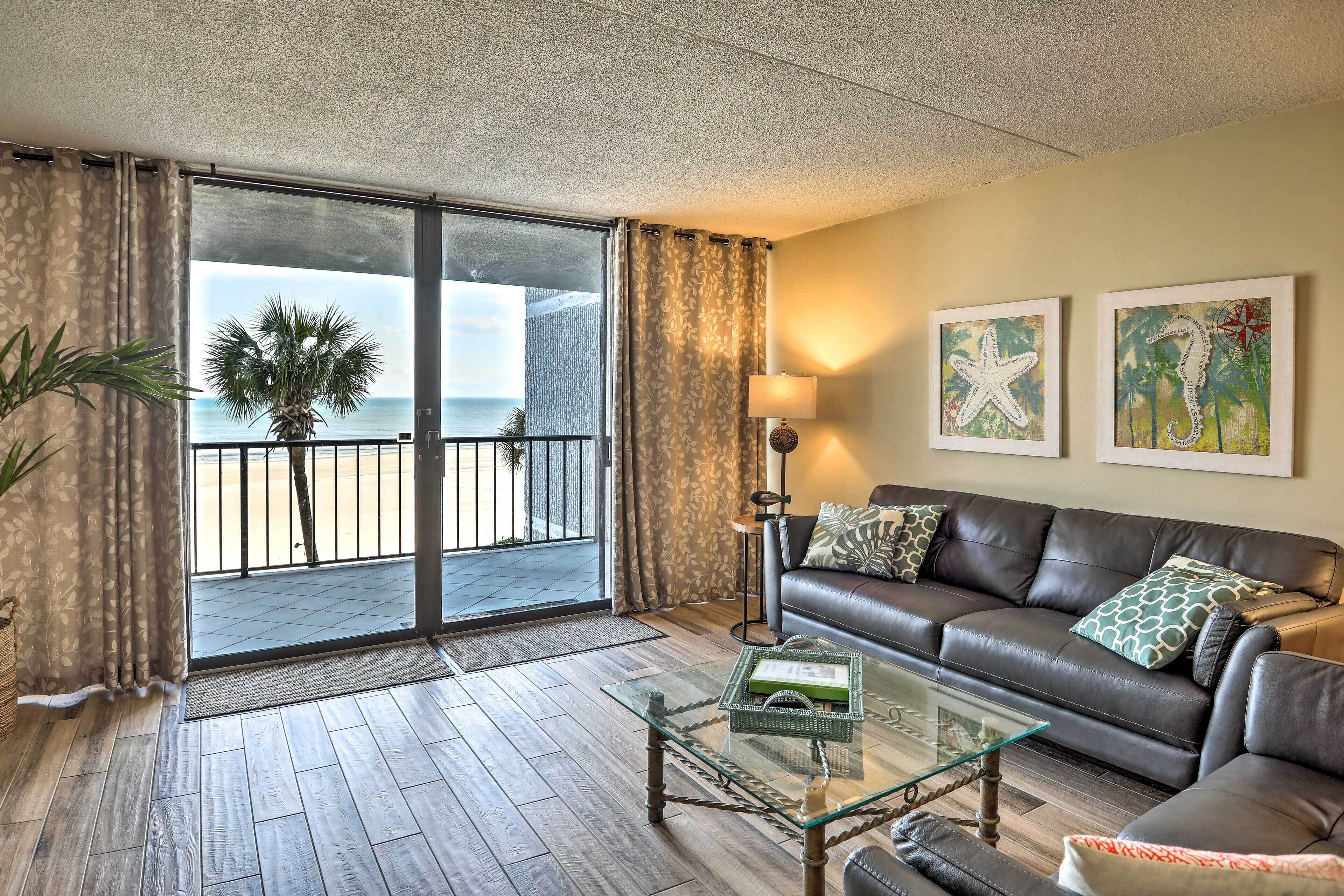Ein Wohnzimmer mit Glastisch, Ledersofa und Blick auf den Strand, sowie einer Tür zu einem Balkon.