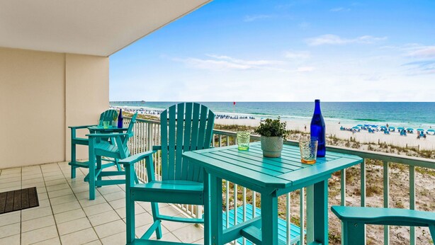 Ein Balkon mit türkisfarbenen Stühlen und einem Tisch, der auf einen Strand mit Menschen und Sonnenschirmen blickt.