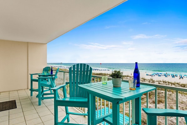 A balcony with turquoise chairs and a table, overlooking a beach with people and umbrellas.