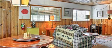 A cozy cabin interior with a wooden dining table, plaid sofa, and a fireplace.