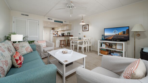 A living room with a teal sofa, white armchairs, a coffee table, and a TV. There's a kitchen area in the background with a dining table and chairs. - Experience your vacation in this spacious one bedroom condo with an ocean view. (Ocean City)