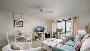 A living room with a white ceiling fan, a television on a white stand, a dining table with chairs, a sofa, and a window with curtains. - Experience your vacation in this spacious one bedroom condo with an ocean view. (Ocean City)