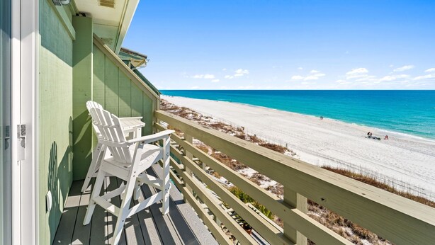 A balcony with white Adirondack chairs overlooking a beach with clear blue water and a sandy shore.