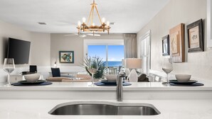 A modern kitchen with a large island, stainless steel sink, and a chandelier. There is a dining area with a table and chairs, and a large window with a view of the ocean.