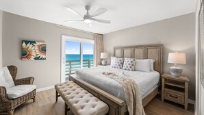 A bedroom with a large bed, a ceiling fan, a view of the ocean, a wicker chair, a bench, and a nightstand with a lamp.