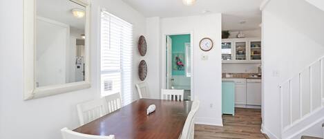 A dining area with a wooden table and white chairs, a large mirror, and a kitchen with white cabinets and a clock.