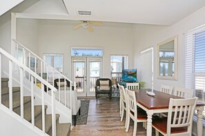 Uma sala de jantar com mesa de madeira e cadeiras brancas, um ventilador de teto e vista para a praia pela porta.