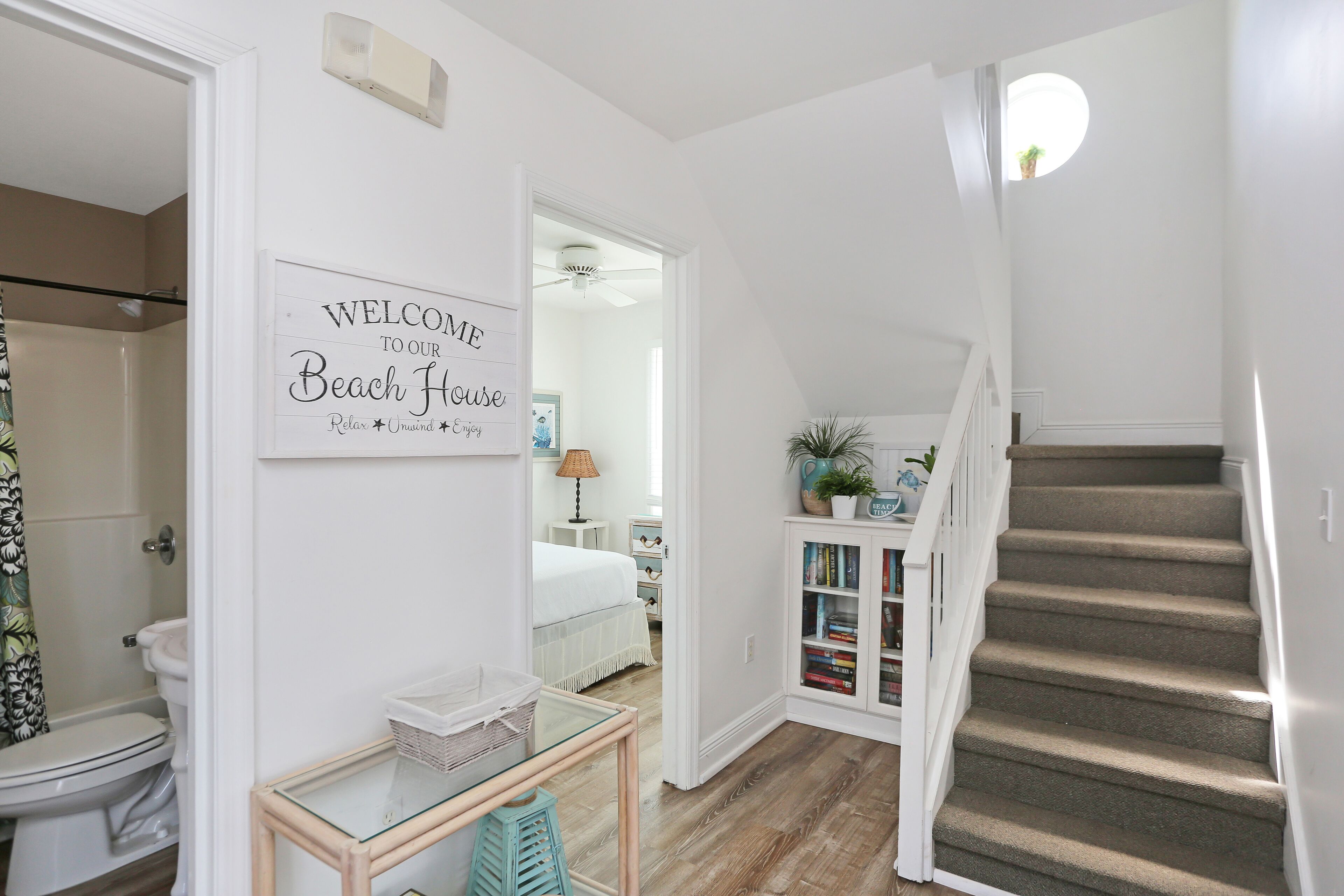 Un couloir avec un escalier, un panneau d’accueil, un lit, une table de chevet et une bibliothèque.