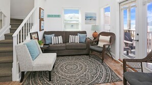 A living room with a grey couch, a grey armchair, a patterned rug, a wicker chair, and a staircase with a sign reading "COASTAL LIVING". - Pet Friendly - Steps From Beach - Narrow Escape (Cape San Blas)