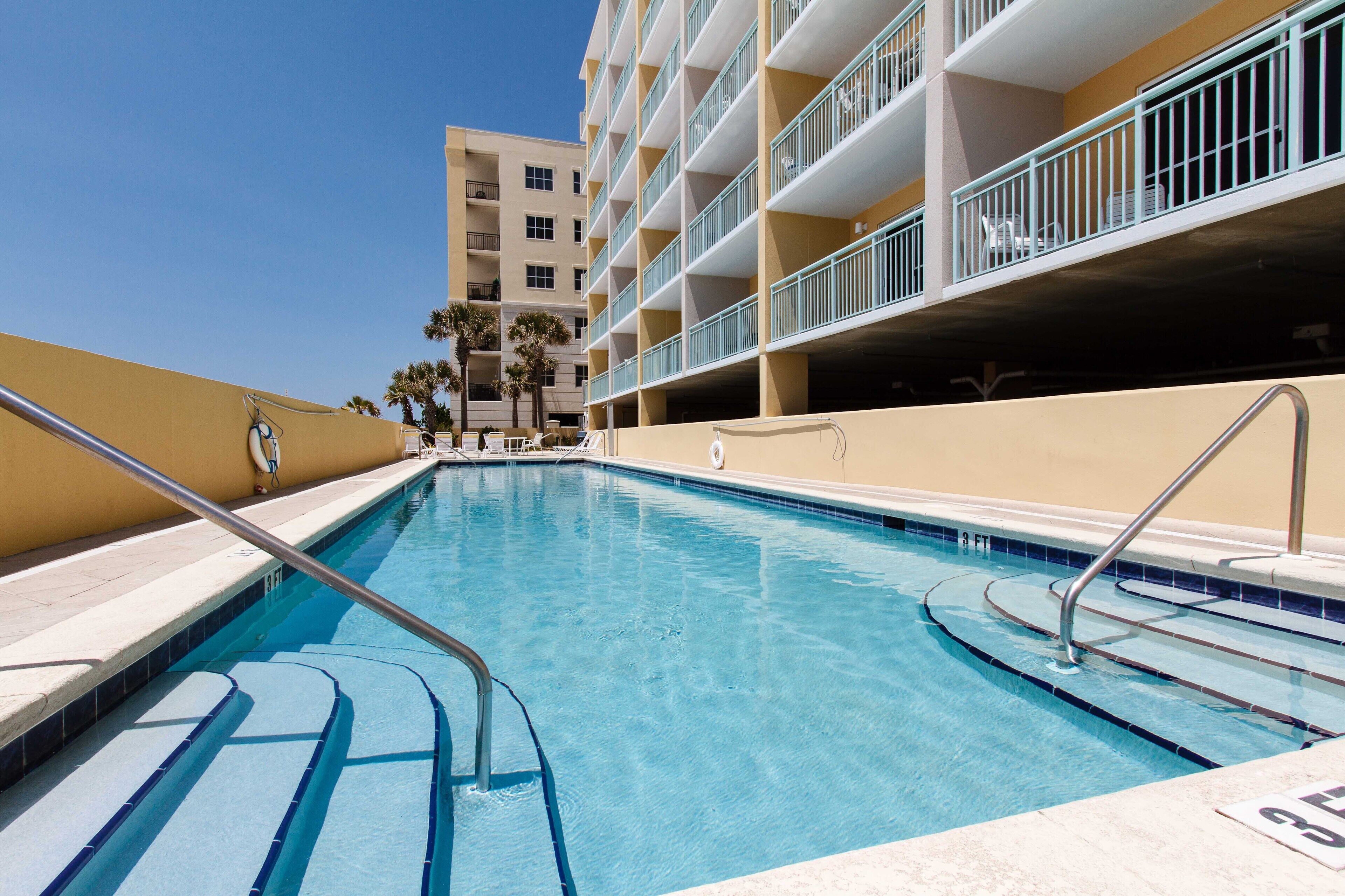 Una piscina con escalones de acceso, rodeada por un edificio con balcones y palmeras.