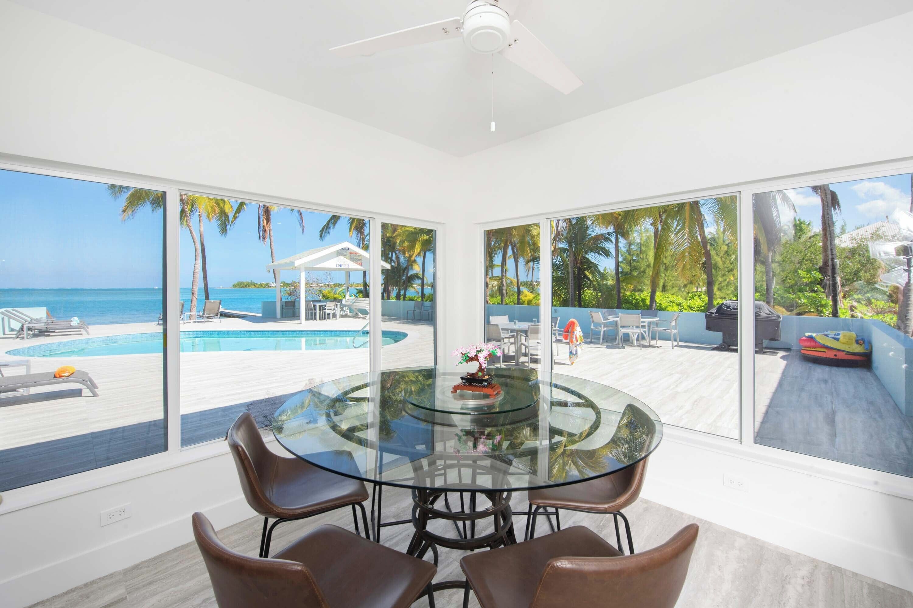 Una habitación moderna con una mesa y sillas de vidrio, ventanas amplias con vista a una piscina y palmeras.