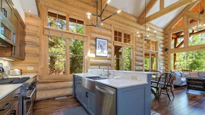 A log cabin kitchen with a central island, large windows, and a dining area with a view of the outdoors. - Lodge-Style Home by InvitedHome | Vast Patio, Hot Tub, Walk to Ski, Fireplace (Mountain Village)