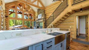 A log cabin interior with a kitchen featuring marble countertops, a stone fireplace, and a staircase. - Lodge-Style Home by InvitedHome | Vast Patio, Hot Tub, Walk to Ski, Fireplace (Mountain Village)
