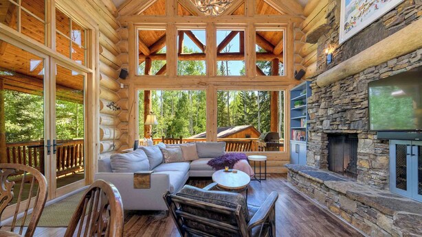 A log cabin interior with a stone fireplace, wooden furniture, and large windows offering a view of the forest. - Lodge-Style Home by InvitedHome | Vast Patio, Hot Tub, Walk to Ski, Fireplace (Mountain Village)