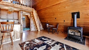 A cozy log cabin interior with a wooden staircase, a kitchen area, a dining table with chairs, and a wood-burning stove. - Secluded & Cozy Antimony Cabin w/ Fire Pit (Antimony)